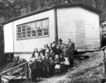 A group of school children of various ages with their male teacher standing in front of a one room white clapboard school house. Photo was taken around 1945.