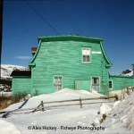 white snow in foreground with a green two-story house occupying the middle ground with a blue sky in the background.
